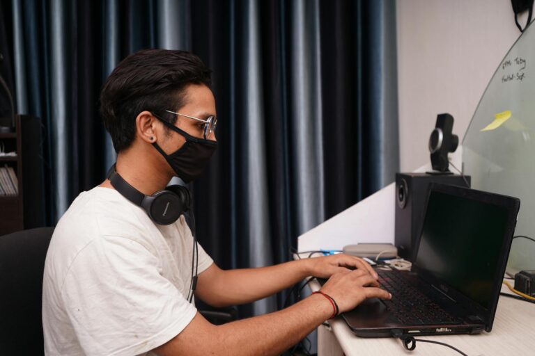 A young man wearing a mask works on his laptop at a home office desk, surrounded by tech accessories.