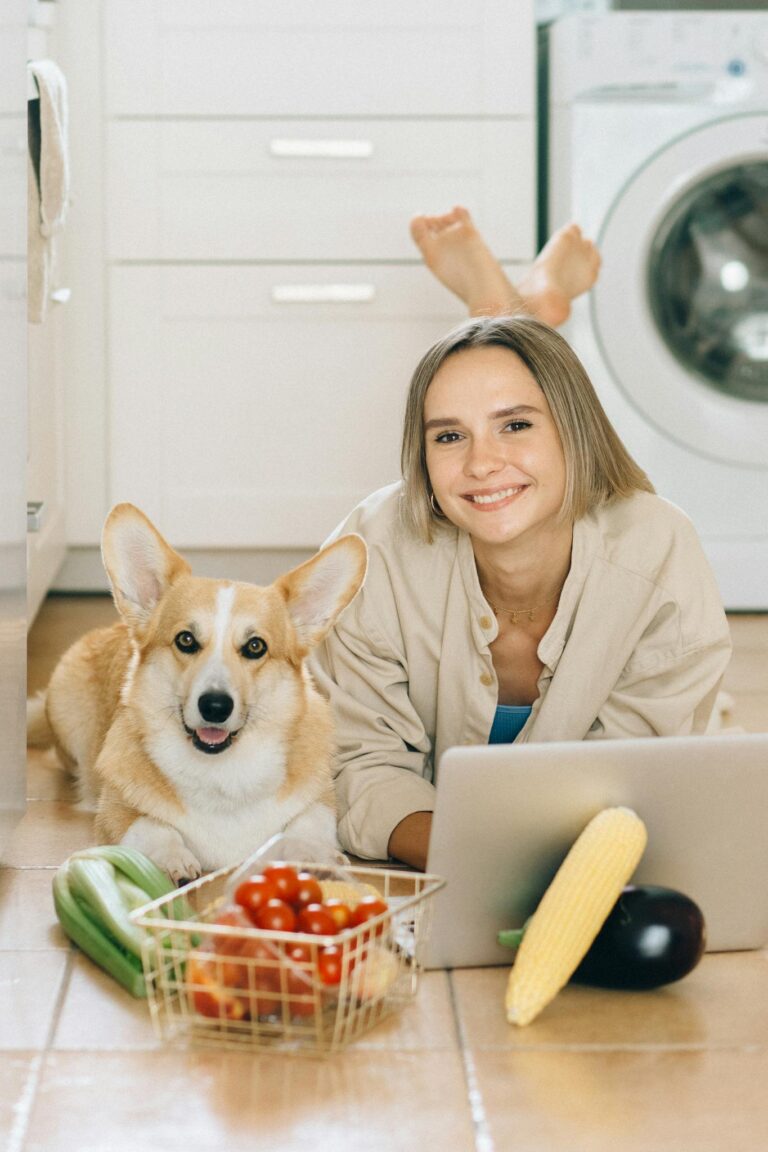 Smiling young woman with a corgi dog, using a laptop in the kitchen surrounded by fresh vegetables.